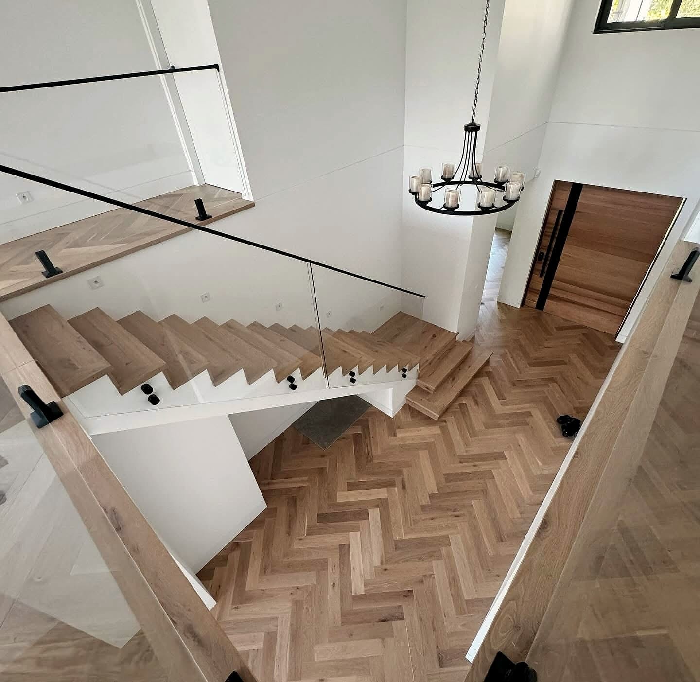 Herringbone floor seen from a landing above, timber stair turning below a chandelier