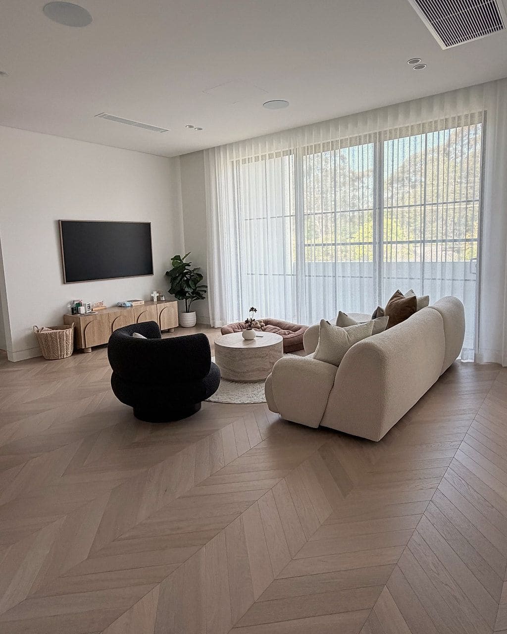 Chevron flooring across an open living room with a curved boucle sofa and a sheer-curtained window wall
