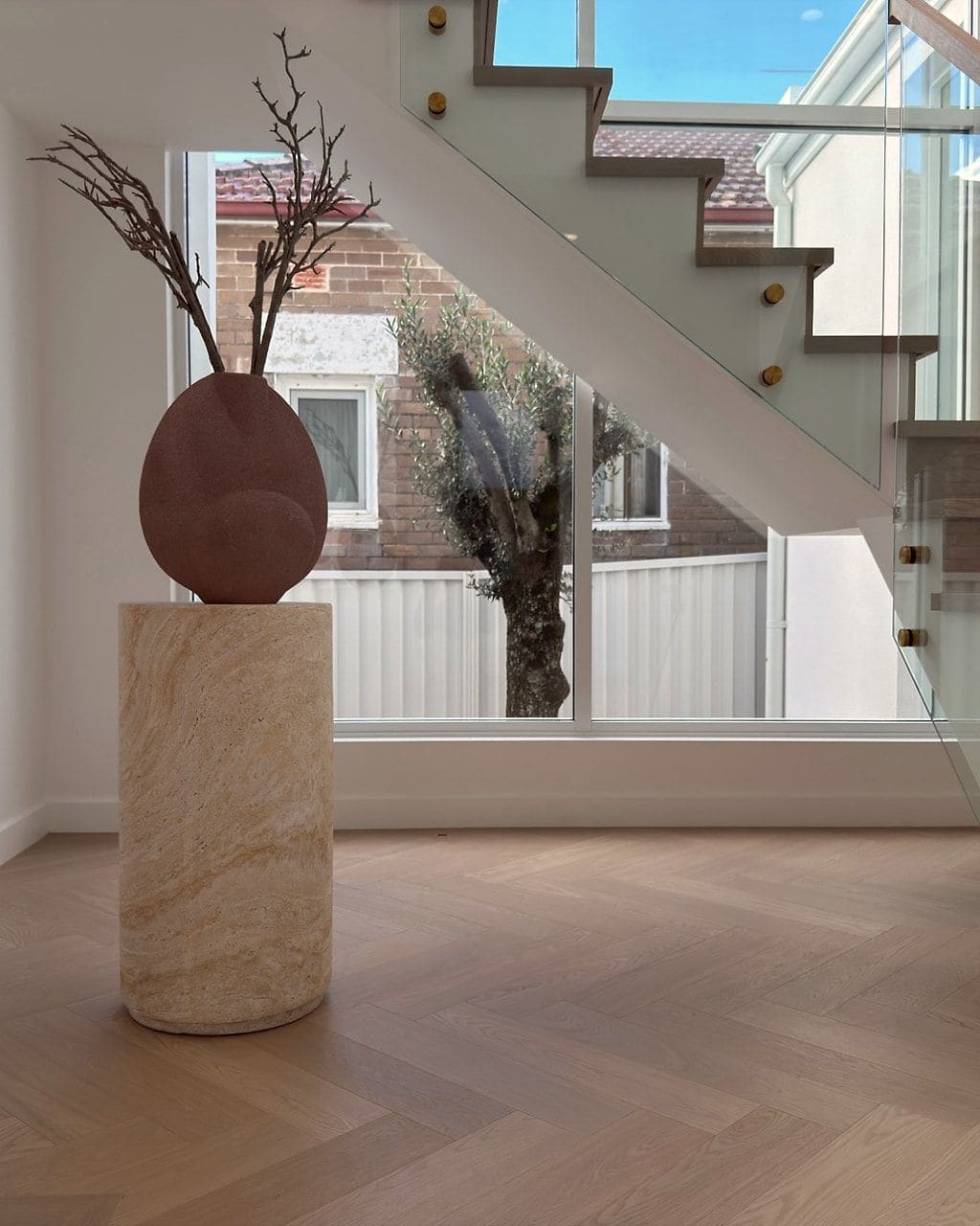 Herringbone oak flooring beneath a glass-balustrade staircase, a stone plinth and sculptural vase in the foreground